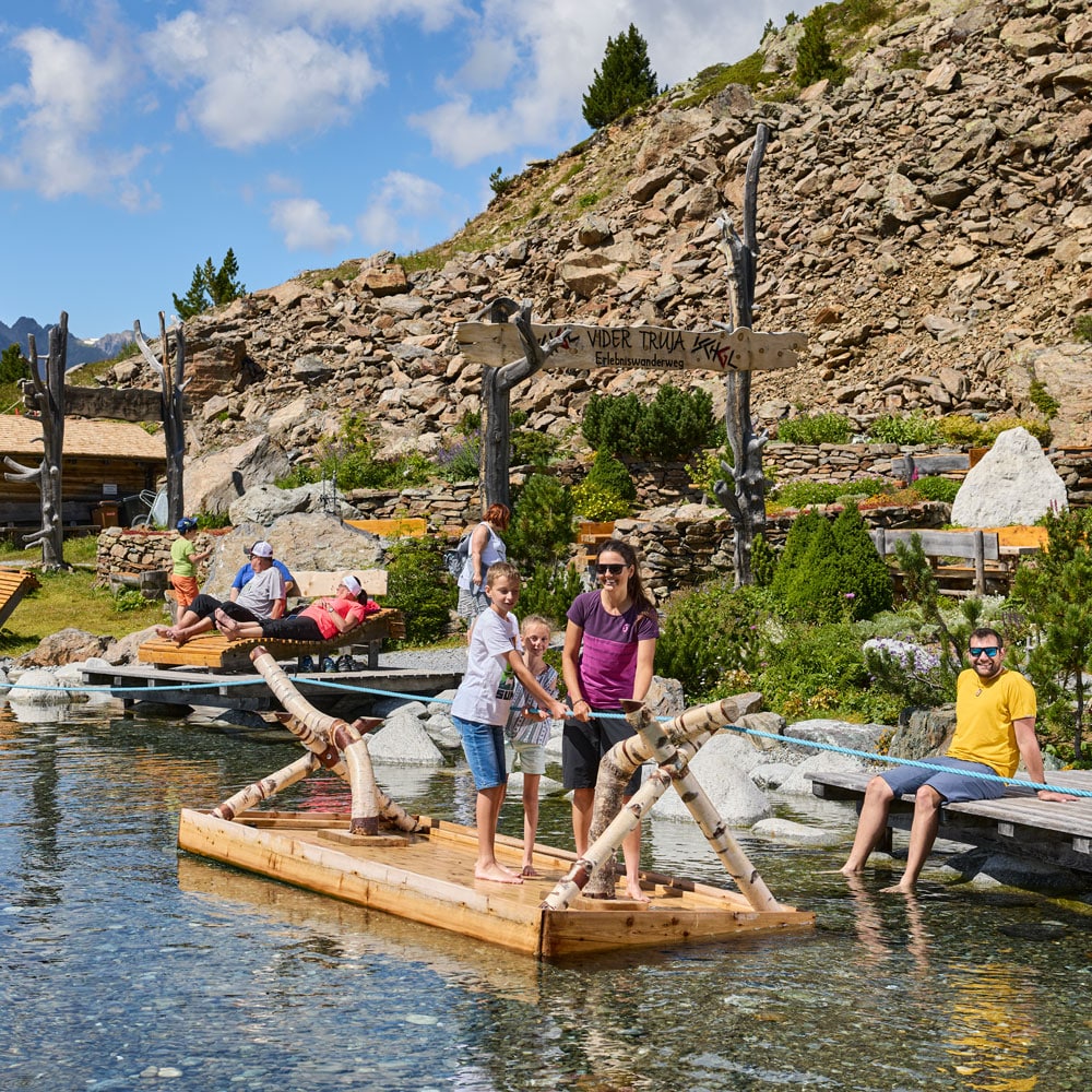 Menschen genießen ein Holzfloß während ihres Sommerurlaubs in Ischgl an einem felsigen Bergteich.