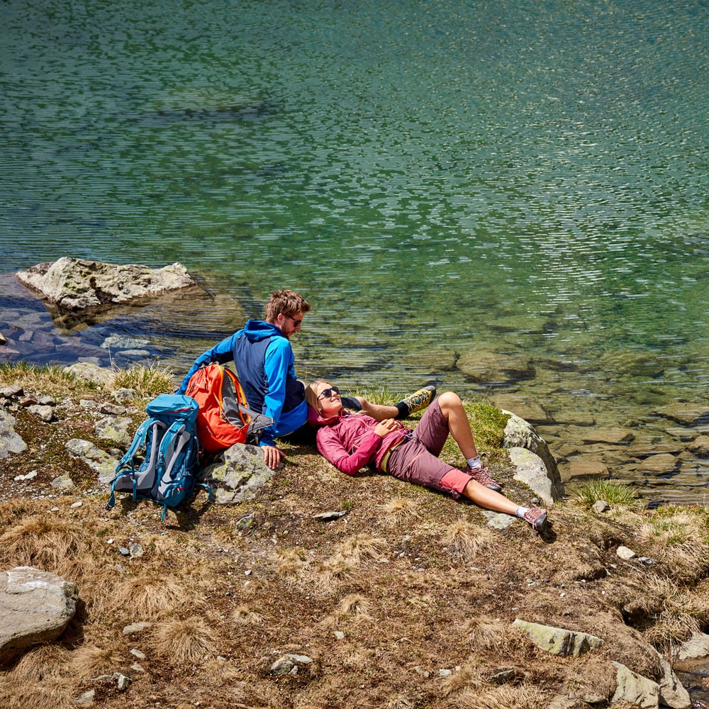 Zwei Personen genießen einen Sommerurlaub in Ischgl an einem klaren Bergsee mit Wanderausrüstung.